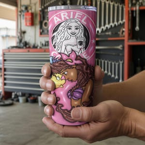May include: A pink tumbler with a cartoon illustration of Ariel and the text "ARIEL" in a circular design. The tumbler also depicts other characters from the movie. The tumbler is held in a person's hands.