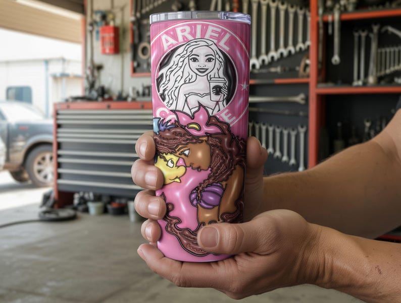 May include: A pink tumbler with a cartoon illustration of Ariel and the text "ARIEL" in a circular design. The tumbler also depicts other characters from the movie. The tumbler is held in a person's hands.