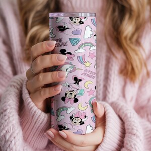 May include: A pink tumbler featuring a repeating pattern of cartoon characters, rainbows, stars, and the text "100% REAL". The tumbler is cylindrical and appears to be made of stainless steel. The design includes Minnie Mouse and other whimsical elements.