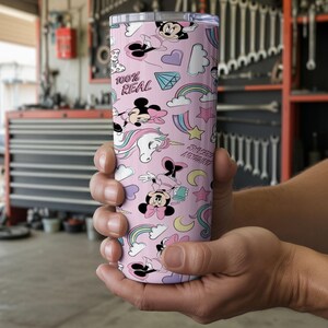 May include: A pink tumbler with a repeating pattern of Minnie Mouse, rainbows, unicorns, and other whimsical elements. The tumbler has the text "100% REAL" and "Sweet Dreams". The tumbler is held in a person's hands.