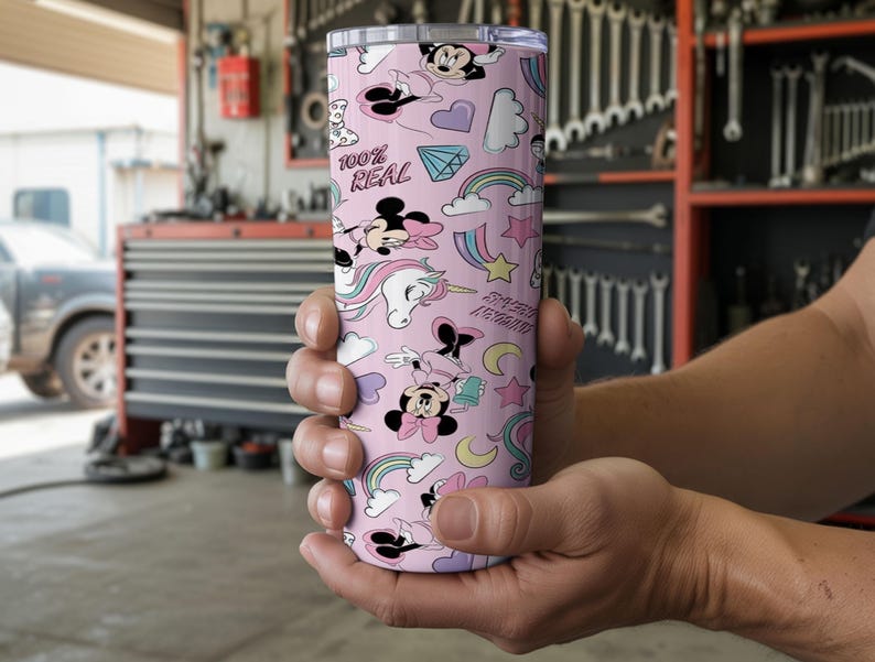 May include: A pink tumbler with a repeating pattern of Minnie Mouse, rainbows, unicorns, and other whimsical elements. The tumbler has the text "100% REAL" and "Sweet Dreams". The tumbler is held in a person's hands.
