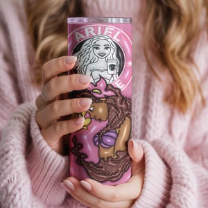 May include: A pink tumbler with a mermaid design and the name "ARIEL". The tumbler features a Starbucks-style logo with a mermaid holding a coffee cup. The bottom of the tumbler has a mermaid tail design.