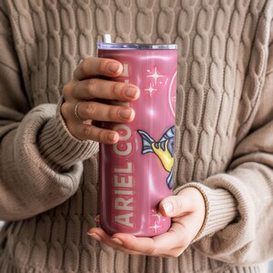 May include: A pink tumbler featuring the text "ARIEL CO." and a cartoon fish illustration. The tumbler has a white straw and a silver lid. The background is a soft, neutral colour.