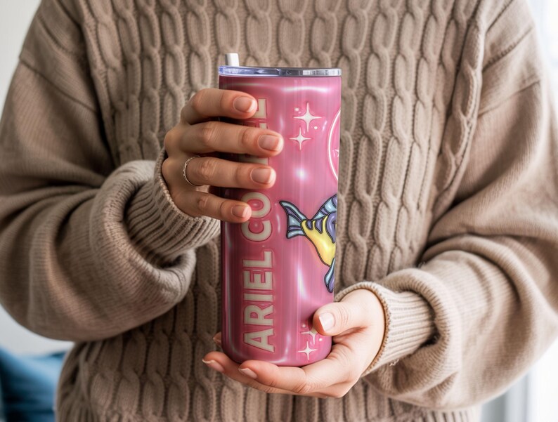 May include: A pink tumbler featuring the text "ARIEL CO." and a cartoon fish illustration. The tumbler has a white straw and a silver lid. The background is a soft, neutral colour.