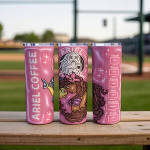 May include: Three pink insulated tumblers with silver lids. One tumbler has "ARIEL COFFEE" in white text and a mermaid illustration. The second tumbler features a mermaid design with the text "ARIEL". The third tumbler has a coffee customization chart.