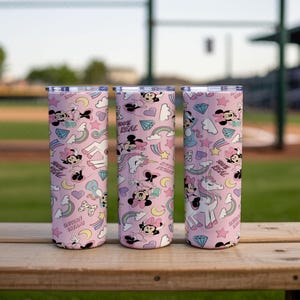 May include: Three pink insulated tumblers with a repeating pattern of Minnie Mouse, rainbows, stars, diamonds, and unicorns. The text "100% REAL" is also visible on the tumblers. The tumblers are cylindrical and have silver lids.