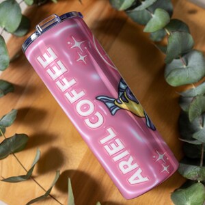 May include: A pink stainless steel travel mug with the words "ARIEL COFFEE" in white, and a cartoon fish illustration. The mug has a silver lid and is surrounded by green eucalyptus leaves.