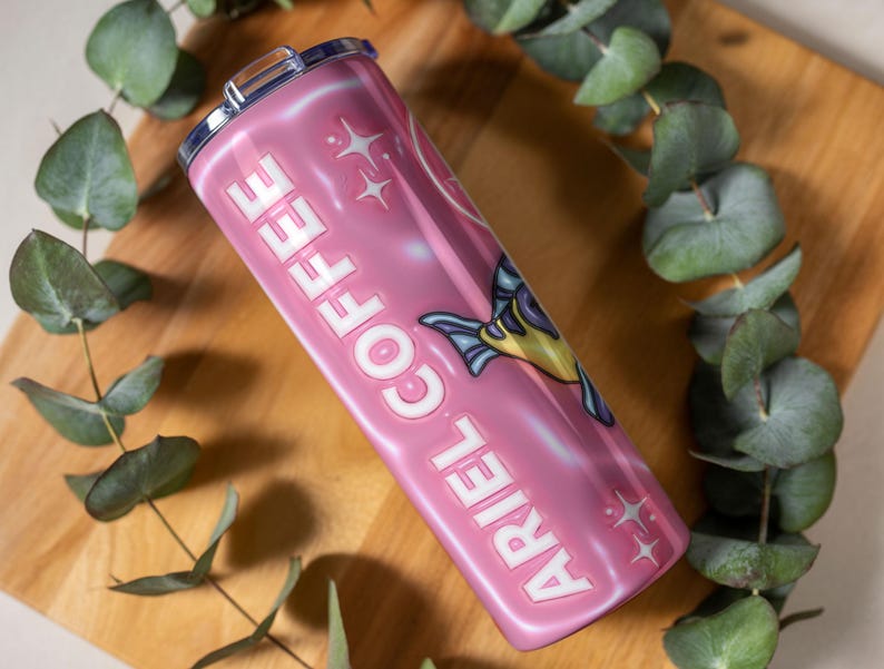 May include: A pink stainless steel travel mug with the words "ARIEL COFFEE" in white, and a cartoon fish illustration. The mug has a silver lid and is surrounded by green eucalyptus leaves.