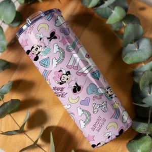 May include: A pink stainless steel tumbler with a lid, featuring a pattern of Minnie Mouse, rainbows, diamonds, stars, and unicorns. The text "100% Real" and "Unicorn Dreams" are also visible on the tumbler.