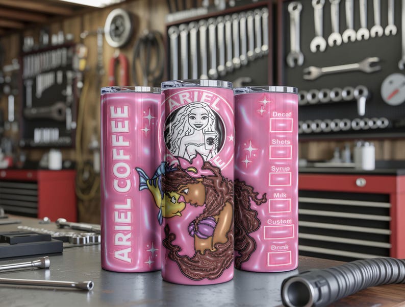 May include: Three pink insulated tumblers with a mermaid theme. One has "ARIEL COFFEE" vertically. Another features a mermaid graphic with the Starbucks logo. The third has a coffee customization chart. The tumblers are on a metal surface.