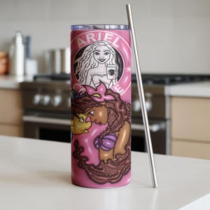 May include: A pink tumbler with a stainless steel straw. The tumbler features a cartoon illustration of Ariel from The Little Mermaid, with the name "ARIEL" in a circular design at the top. The design includes Ariel holding a coffee cup.