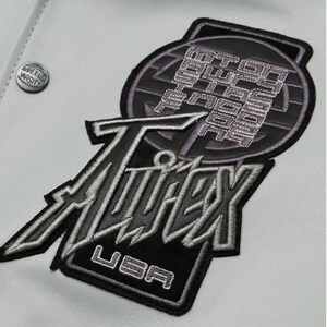 May include: Close-up of a white jacket with embroidered patches. The patches feature the text "Twitex USA" in silver and black, and a circular patch with text. A small "Apex USA Varsity" button is visible.