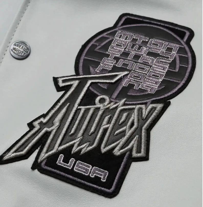 May include: Close-up of a white jacket with embroidered patches. The patches feature the text "Twitex USA" in silver and black, and a circular patch with text. A small "Apex USA Varsity" button is visible.