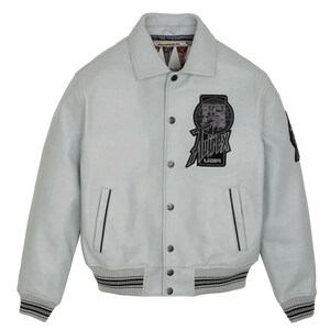 May include: A light gray leather jacket with a button-up front, featuring a black and white logo patch on the chest that reads "Avirex USA." The jacket has a ribbed hem and cuffs with black and white stripes, and a collar.