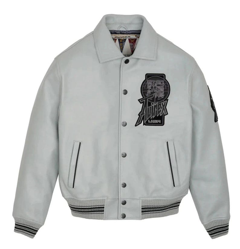 May include: A light gray leather jacket with a button-up front, featuring a black and white logo patch on the chest that reads "Avirex USA." The jacket has a ribbed hem and cuffs with black and white stripes, and a collar.