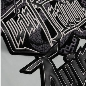 May include: Close-up of a black and gray patch with white trim. The patch features stylized lettering and a dragon design. The background is a light gray color.