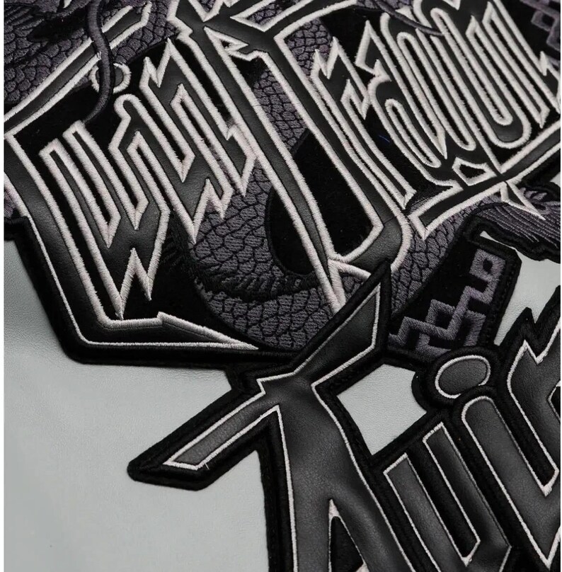 May include: Close-up of a black and gray patch with white trim. The patch features stylized lettering and a dragon design. The background is a light gray color.