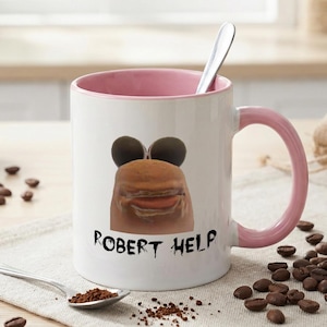 Puede incluir: Taza de cerámica blanca con interior y asa rosas, con una cara de dibujos animados y el texto "ROBERT HELP" en negro. Una cuchara plateada descansa sobre una superficie blanca con granos de café esparcidos.