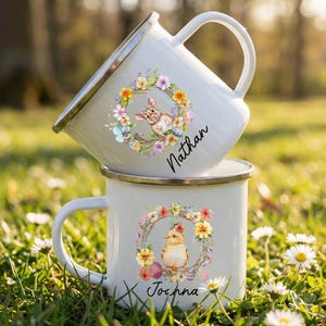 May include: Two white enamel mugs with silver rims are stacked on top of each other. The top mug features a bunny in a floral wreath and the name "Nathan". The bottom mug has a chick in a floral wreath and the name "Jochna".