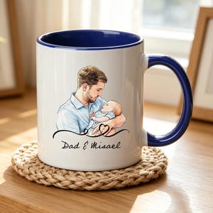 Op de afbeelding: Witte keramische mok met een blauwe binnenkant en handvat. De mok heeft een illustratie van een vader die een baby vasthoudt, met de tekst "Dad & Misael" eronder. De mok staat op een ronde, geweven onderzetter.