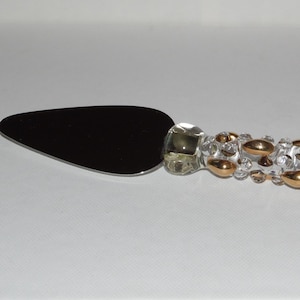 May include: A cake server with a black blade and a clear handle adorned with gold and clear decorative elements. The handle has a twisted gold accent at the end, adding a touch of elegance to this serving utensil.