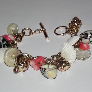 May include: A charm bracelet featuring a rose gold-toned chain with a toggle clasp. The bracelet is adorned with various natural elements, including white ridged seashells, spiral shells, speckled black and white shells, and smooth pink coral-like pieces.