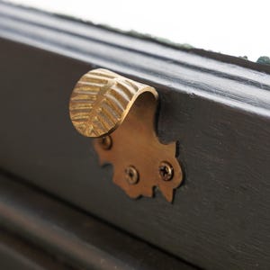May include: A brass drawer pull with a curved handle and decorative backplate. The handle has a textured, ridged design. The backplate is attached with two screws to a dark wooden surface.