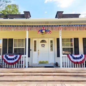 May include: A house decorated with patriotic bunting and an eagle emblem. The porch features red, white, and blue decorations, including a banner with stars and stripes. The house number "555" is visible on a white column.