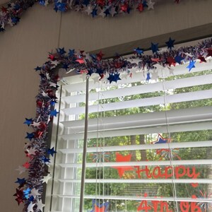 May include: A festive garland of red, white, and blue star-shaped decorations adorns a window. The garland is draped across the top of the window and along the side, creating a patriotic display. The window blinds are white, and the text "Happy 4th of July" is visible.