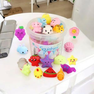May include: A collection of colorful squishy toys in various shapes, including animals, fruits, and stars. The toys are in a clear plastic container with the word "Squishies" printed on it. The toys are made of soft, squeezable material.