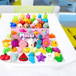 May include: A collection of colorful mochi squishies in various shapes, including animals, fruits, and flowers. The squishies are arranged on a white tray, with a pink box labeled "Mochi Squishy" in the background.
