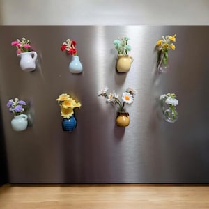 May include: A collection of small, decorative vases, each filled with colorful artificial flowers, are attached to a stainless steel surface. The vases come in various shapes and colors, including white, blue, yellow, and brown, creating a charming display.