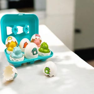 May include: A teal egg-shaped container holding six white eggs with dinosaur designs. Each egg contains a different colored dinosaur toy. One egg is cracked open, and another egg is separate from the container.