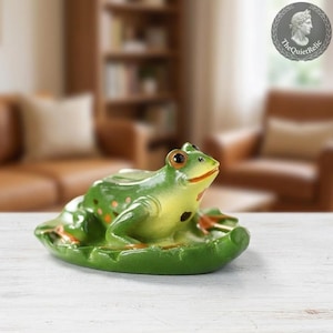 May include: A ceramic frog figurine in shades of green and yellow, with orange spots, resting on a green lily pad. The frog has large, dark eyes and a smiling red mouth. The figurine is a decorative item.