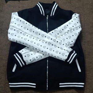 Könnte beinhalten: Schwarze Collegejacke mit weiß gemusterten Ärmeln. Die Jacke hat einen durchgehenden Reißverschluss, weiße Streifen an den Bündchen und am Saum sowie einen weißen Kragen. Die Ärmel zeigen ein sich wiederholendes Muster aus Diamanten und Buchstaben.