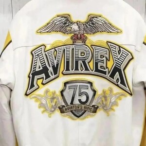 Puede incluir: Chaqueta bomber blanca y amarilla con la palabra "AVIREX" en letras grandes, negras y amarillas. La chaqueta presenta un emblema de &aacute;guila y el texto "75 AVIATOR'S KING".