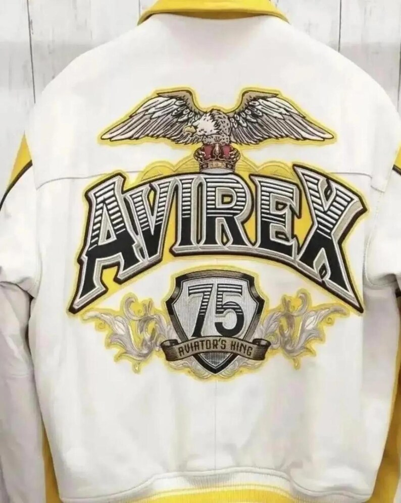 Puede incluir: Chaqueta bomber blanca y amarilla con la palabra "AVIREX" en letras grandes, negras y amarillas. La chaqueta presenta un emblema de &aacute;guila y el texto "75 AVIATOR'S KING".