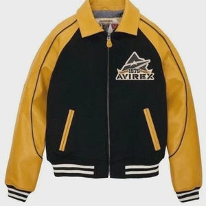 Puede incluir: Chaqueta bomber negra y amarilla con cremallera. La chaqueta presenta mangas y cuello de cuero amarillo, con un cuerpo negro. La parte delantera muestra un logotipo blanco y amarillo con el texto "BOSTON 1979 CLASSIC AVIREX". La chaqueta tiene dobladillo y pu&ntilde;os acanalados.