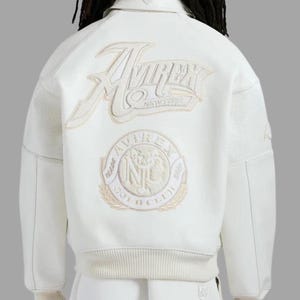May include: Cream-colored jacket with embroidered logos. The back features a large "AVIREX NEW YORK" logo and a circular "GOLD CLUB" emblem. The jacket has perforated panels on the shoulders and ribbed detailing at the waist.