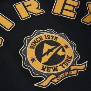 Puede incluir: Tela negra con un parche bordado amarillo y blanco. El parche dice "SINCE 1975 NEW YORK CLASSIC" alrededor de un logotipo central. Se ve texto amarillo y blanco adicional en la parte superior.