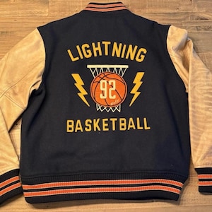 Puede incluir: Chaqueta universitaria azul marino y beige con el texto "LIGHTNING BASKETBALL 92". La chaqueta tiene mangas de cuero beige, cuerpo azul marino y ribete a rayas naranja y blanco en los puños y el cuello.