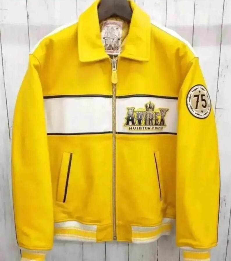 Puede incluir: Chaqueta de cuero amarilla con una franja blanca en el pecho y las mangas. Presenta el logotipo "AVIREX AVIATOR'S KING" y un parche "75". Incluye cierre de cremallera y cuello.