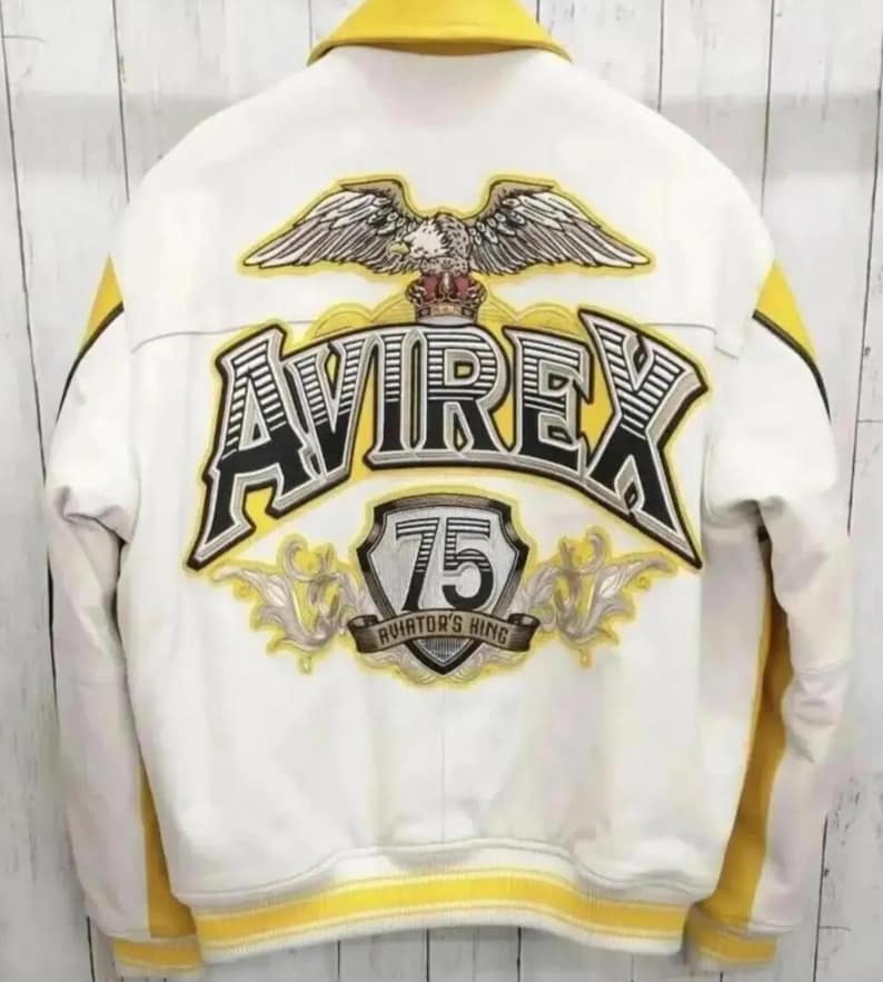 Puede incluir: Chaqueta de cuero blanca con detalles amarillos y cuello amarillo. La parte trasera presenta un gran logotipo "AVIREX", un emblema de &aacute;guila y el n&uacute;mero "75" con el texto "AVIATOR'S KING". La chaqueta tiene un estilo bomber cl&aacute;sico.
