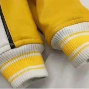 Puede incluir: Primer plano de una chaqueta universitaria amarilla y blanca. La chaqueta presenta mangas amarillas y un cuerpo blanco con una raya negra. Los pu&ntilde;os son blancos con rayas amarillas. La chaqueta es de material suave.