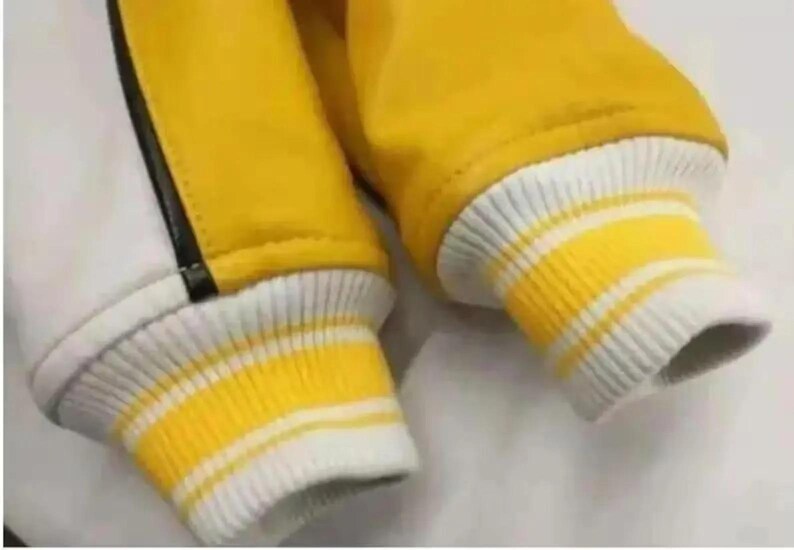Puede incluir: Primer plano de una chaqueta universitaria amarilla y blanca. La chaqueta presenta mangas amarillas y un cuerpo blanco con una raya negra. Los pu&ntilde;os son blancos con rayas amarillas. La chaqueta es de material suave.