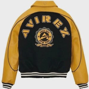 Puede incluir: Una chaqueta universitaria negra y amarilla con el nombre de la marca "AVIREX" en letras amarillas. La parte trasera de la chaqueta presenta un emblema circular con el texto "SINCE 1975 NEW YORK CLASSIC". La chaqueta tiene un dobladillo acanalado con rayas blancas y negras.