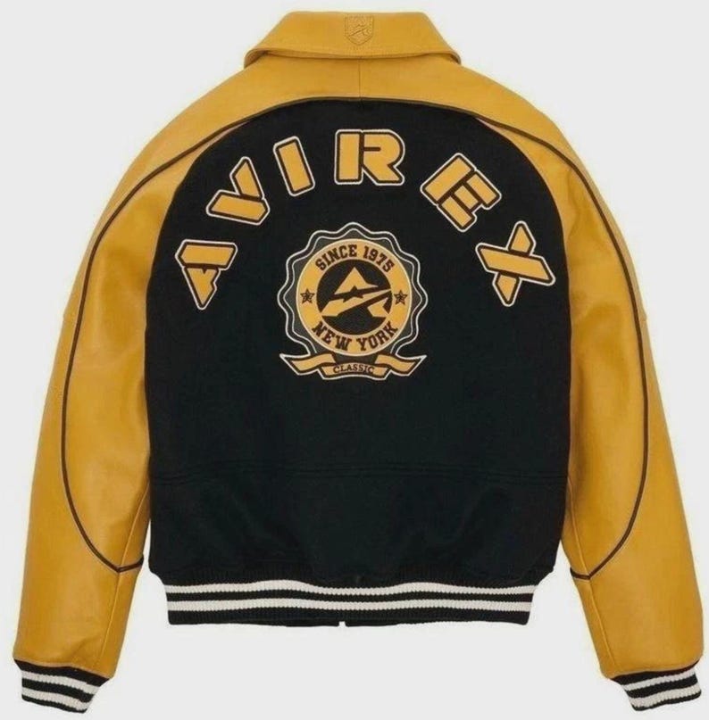 Puede incluir: Una chaqueta universitaria negra y amarilla con el nombre de la marca "AVIREX" en letras amarillas. La parte trasera de la chaqueta presenta un emblema circular con el texto "SINCE 1975 NEW YORK CLASSIC". La chaqueta tiene un dobladillo acanalado con rayas blancas y negras.