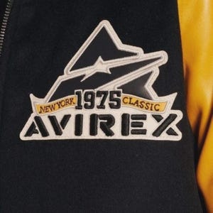 Puede incluir: Primer plano de una chaqueta negra y amarilla con un parche. El parche dice "NEW YORK 1975 CLASSIC AVIREX" y presenta un dise&ntilde;o de monta&ntilde;a en blanco y negro con una pancarta amarilla.