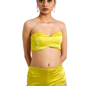 Peut inclure: Ensemble composé d'un haut bustier en satin jaune et d'un short assorti. Le haut a une encolure en cœur et le short est taille haute. La tenue est d'une couleur unie vibrante et le tissu a un léger éclat. Le modèle est devant un fond blanc.