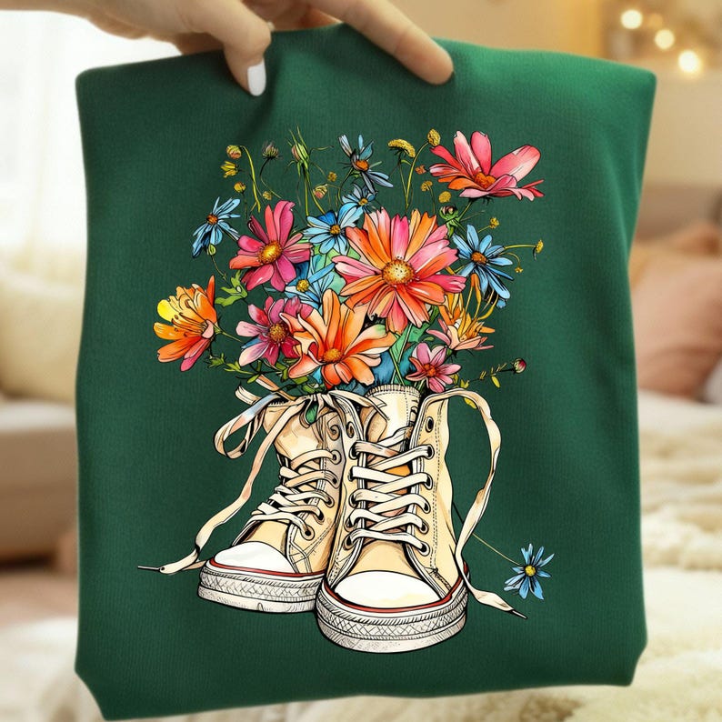 May include: A dark green sweatshirt featuring a colorful floral design. The graphic depicts a pair of cream-colored high-top sneakers filled with a bouquet of vibrant flowers in shades of pink, orange, blue, and yellow.